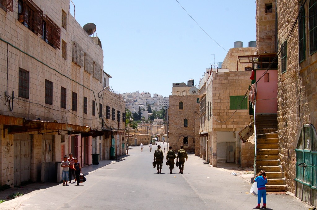 Israeli soldiers walk down the center of the deserted Shuhada Street as Palestinian children linger on the sides.  